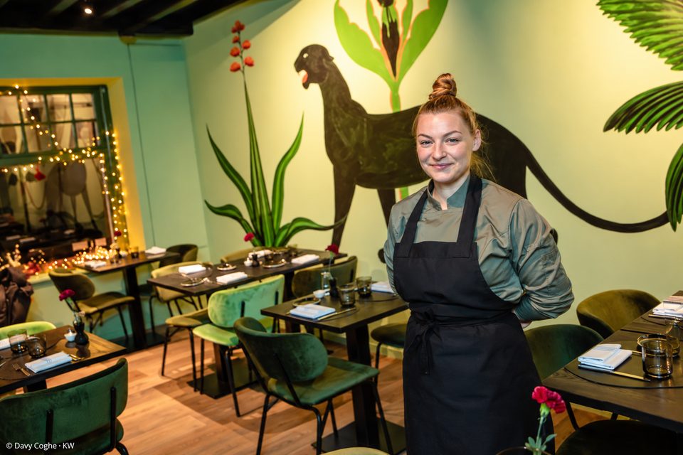 Louise Fonteyne in koksjas en zwarte schort, lachend in het restaurant, met de panterschildering op de muur en de groene fluwelen stoelen achter haar
